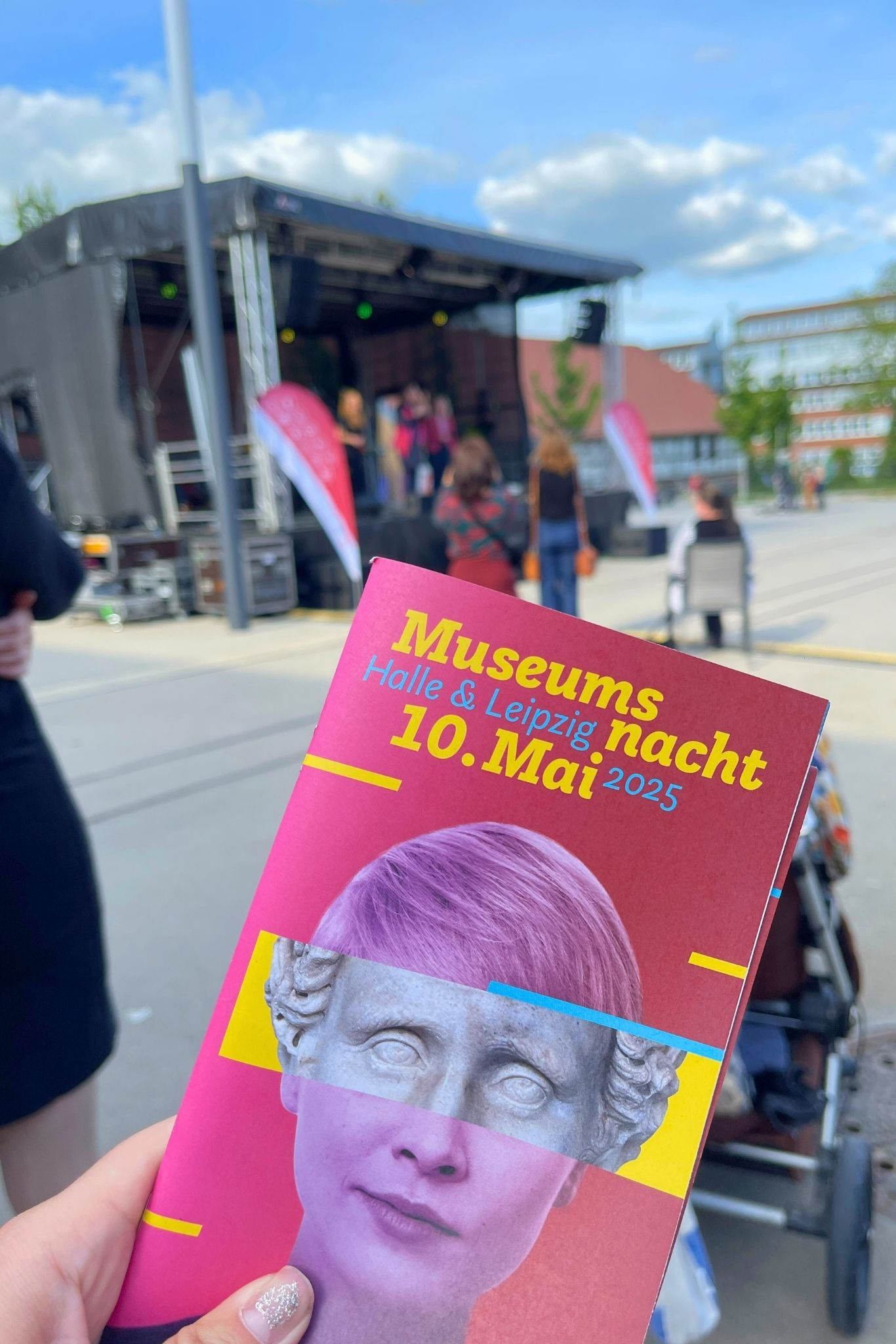 Eine Person hält ein farbiges Veranstaltungsfaltblatt mit dem Titel „Museumsnacht Halle & Leipzig 10. Mai 2025“ in die Kamera. Im Hintergrund eine Bühne mit Technik und Publikum auf dem Salinehof.