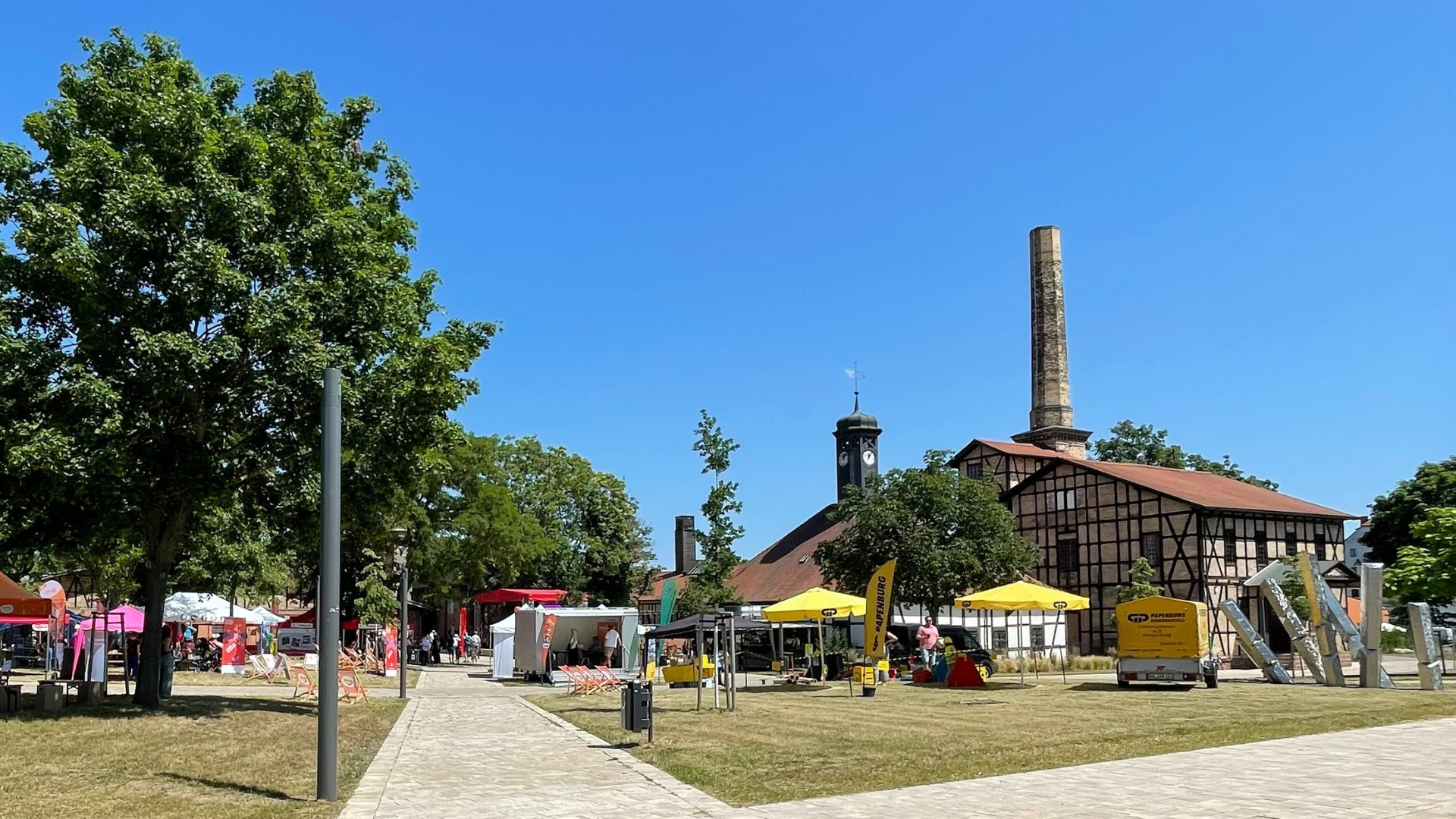 Außengelände des Salinemuseums. Rechts im Hintergrund das Uhrenhaus. Im Vordergrund auf der Wiese und dem gepflasterten Platz sind verschiedene Pavillons und Stände aufgebaut. Besucher:innen bewegen sich zwischen den Zelten. Es ist schönes Wetter. Der Himmel ist strahlend blau.